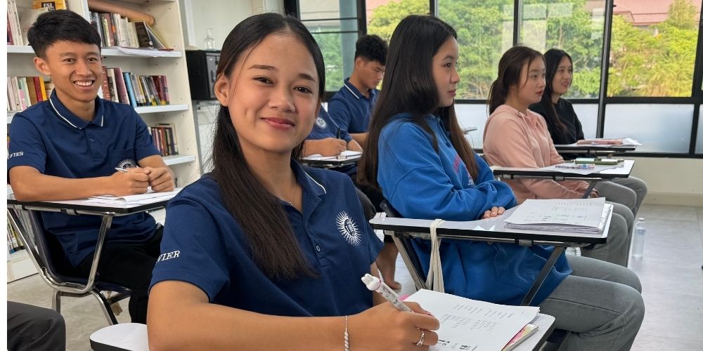 Fa smiling as she writes in her notebook in a brightly-lit classroom surrounded by her fellow students