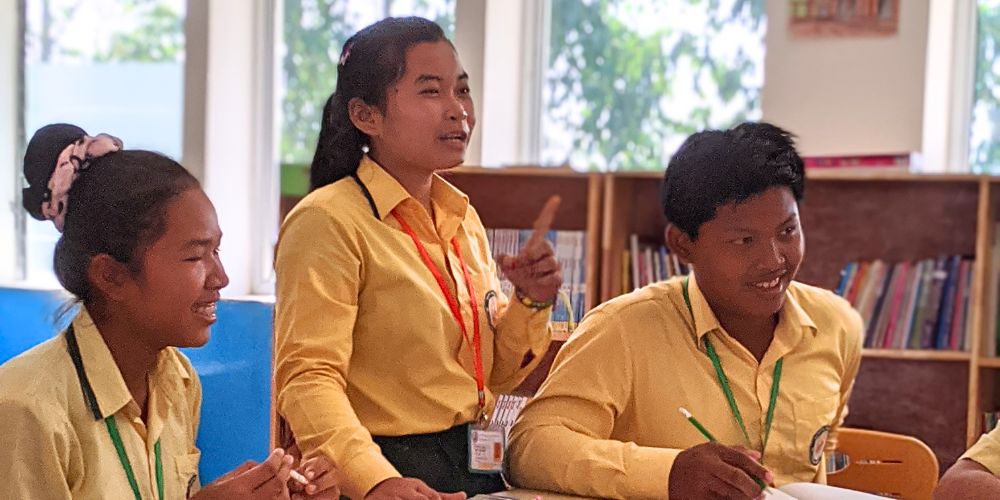 Kanya answering a question in class at a desk with two of her classmates