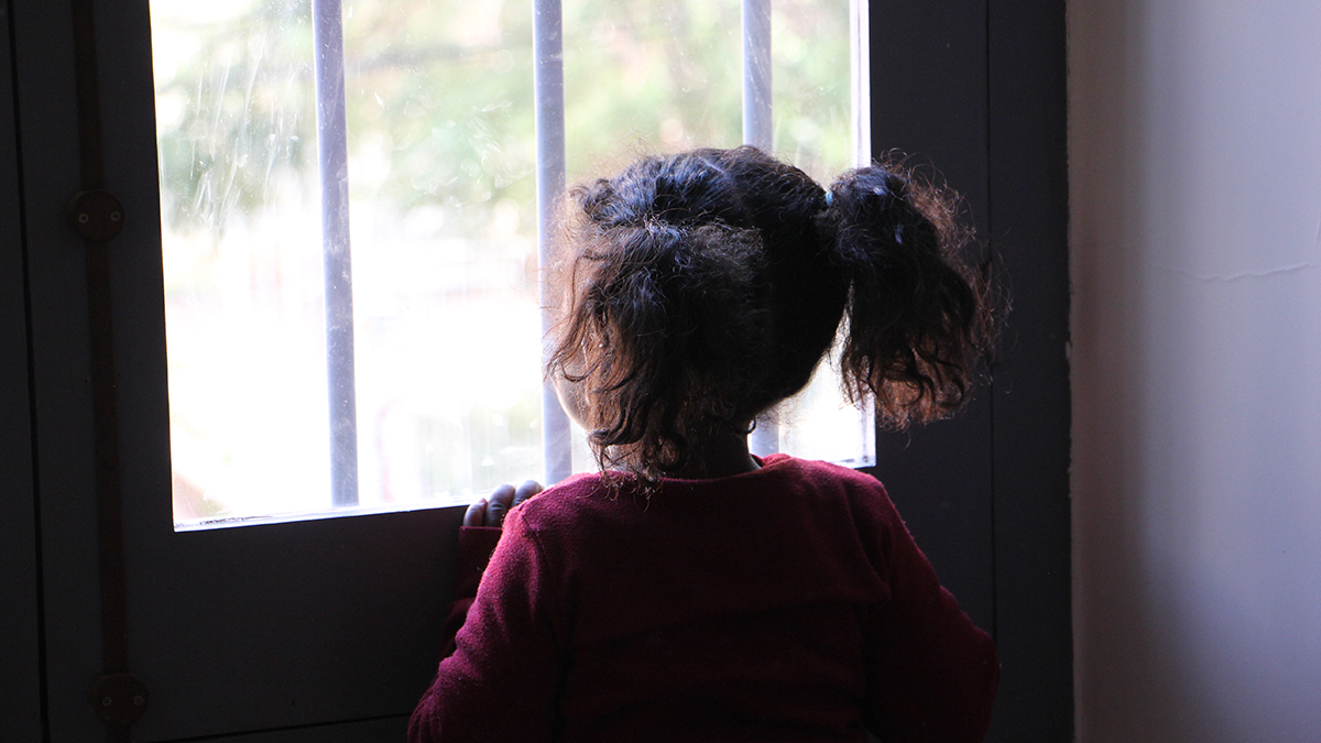 A young child looks longingly out a window from a darkened room