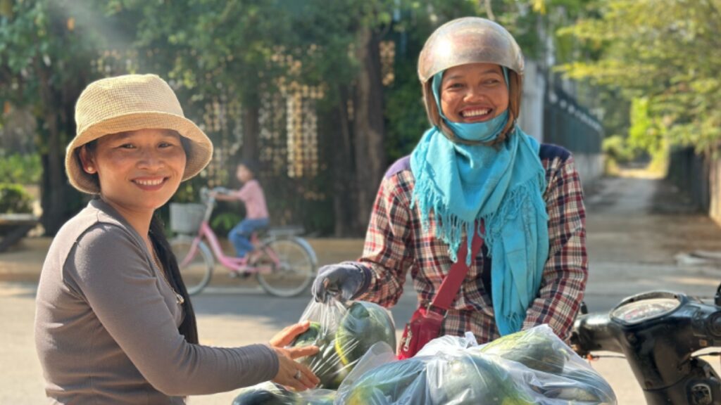 Tut Ngah selling fruit from the back of her motorbike to a woman from her community