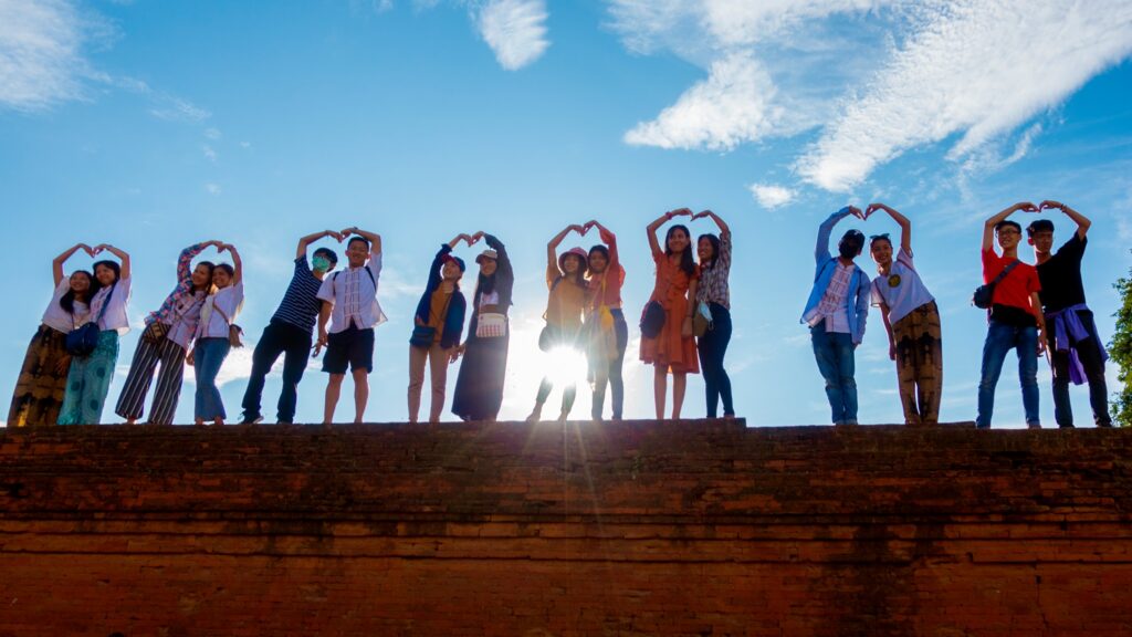 Young people lined up on a wall making love hearts with their arms with the sun shining behind them