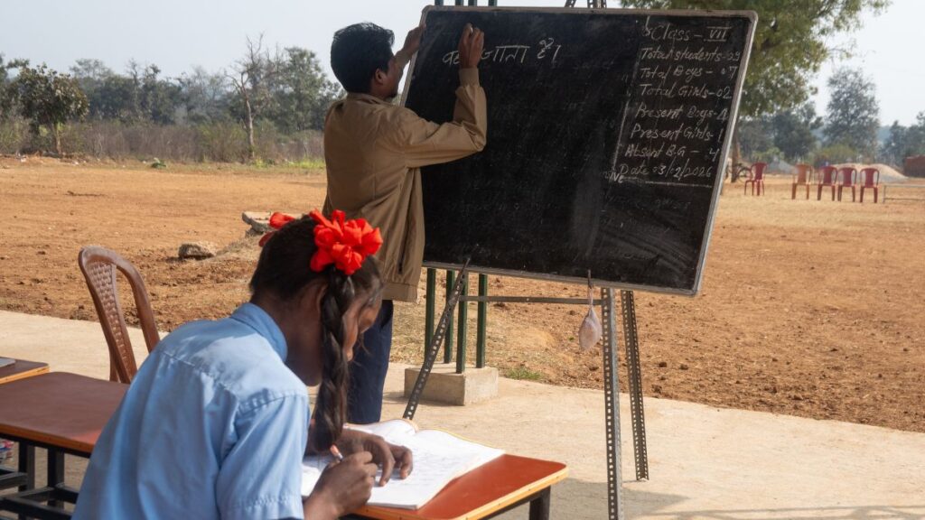 A teacher writing on a mobile blackboard while a student takes notes. The class is outside in the sunshine